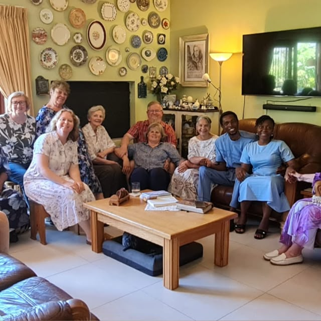 John and Grace Damilola with brothers and sisters gathered around a dining table in a living room with decorative plates on the wall, smiling together